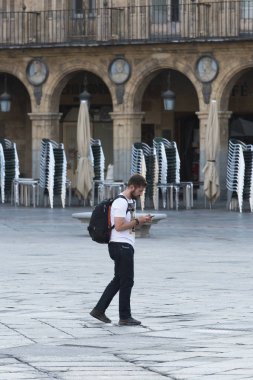 Salamanca, İspanya - 2 Eylül 2017: Sırt çantalı genç bir adam Plaza Mayor 'da yürürken cep telefonunu kontrol ediyor.