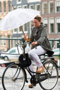 AMSTERDAM, NETHERLANDS - 8 HAZİRAN 2015: Bisiklet süren ve şemsiye taşıyan bir kadın, şehrin bir caddesinde sağanak yağmur altında yürüyor.