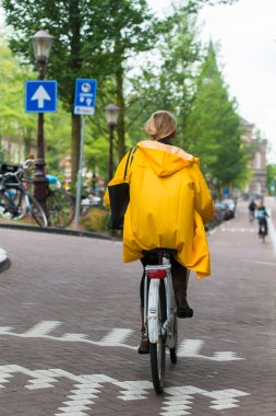 AMSTERDAM, NETHERLANDS - 8 Temmuz 2015: Gösterişli sarı yağmurluklu bir kadın, şehrin sokaklarından birinde bisiklet sürüyor.