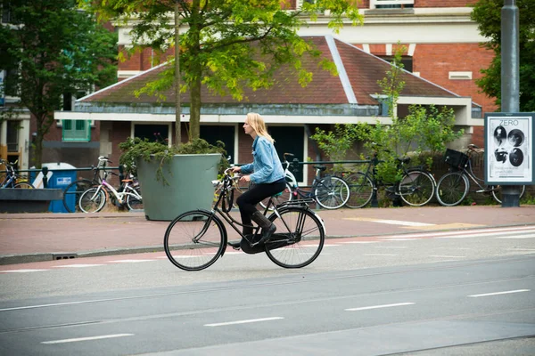 AMSTERDAM, NETHERLANDS - 8 HAZİRAN 2015: Siyah bisikletli genç bir sarışın şehrin sokaklarından birinin yanından geçiyor.
