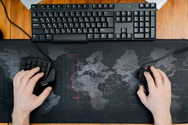 one hand on the gaming keyboard, the second on a computer mouse on a background of a computer mat with a world map