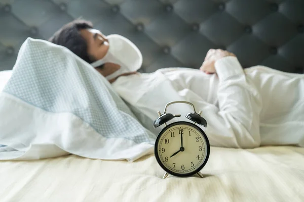 Asian businessman wearing surgical mask sleeping on the bed with alarm clock