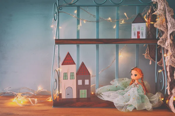 Cute doll and wooden little houses next to warm garland lights in front of blue background.