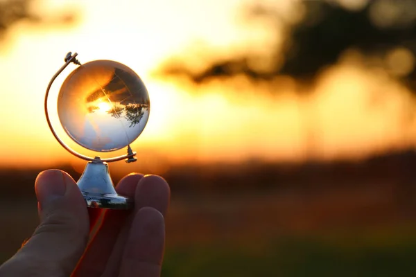 Mano masculina sosteniendo pequeño globo de cristal delante de la puesta del sol — Foto de Stock