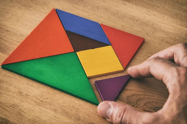 man's hand holding a missing piece in a square tangram puzzle, over wooden table