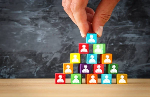 Main de l'homme tenant un sommet de la pyramide de blocs de bois avec des icônes de personnes sur la table en bois — Photo