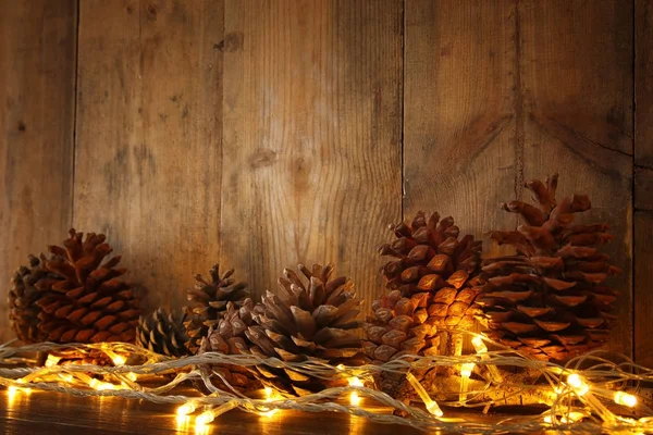 Holiday image with Christmas golden garland lights and pine cones over wooden background — Stock Photo, Image