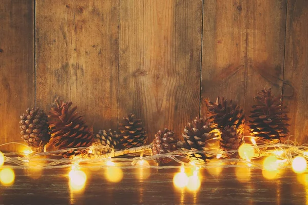 Imagen navideña con luces de guirnalda dorada y conos de pino sobre fondo de madera — Foto de Stock