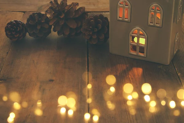 Low key image of house lantern with burning candle and warm light in the windows over old wooden background.