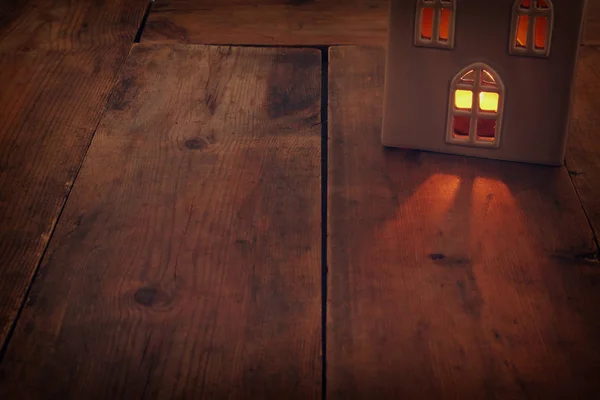 Low key image of house lantern with burning candle and warm light in the windows over old wooden background.