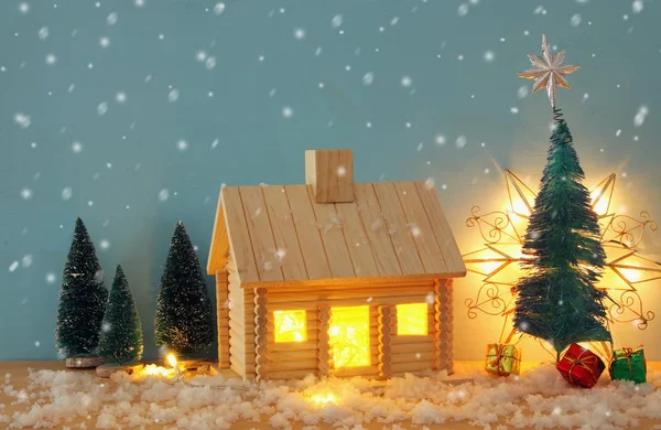 Image de sapin de Noël et maison en bois avec lumière à travers la fenêtre, sur une table enneigée . — Photo