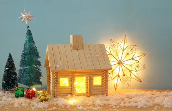 Image de sapin de Noël et maison en bois avec lumière à travers la fenêtre, sur une table enneigée . — Photo