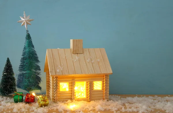 Afbeelding van de kerstboom en houten huis met licht door het raam, boven besneeuwde tafel. — Stockfoto