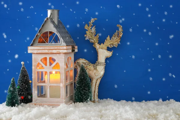 木制老房子与蜡烛, 白色鹿在雪和蓝色 nackground 旁边在圣诞节树. 免版税图库照片