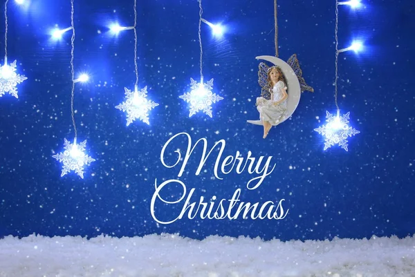Magical christmas image of little white fairy with glitter wings sitting on the moon over blue background and silver snowflake garland. — Stock Photo, Image