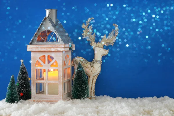 Деревянный старый дом со свечой, белый олень рядом с рождественскими деревьями на снегу и голубой настил . Стоковое Изображение