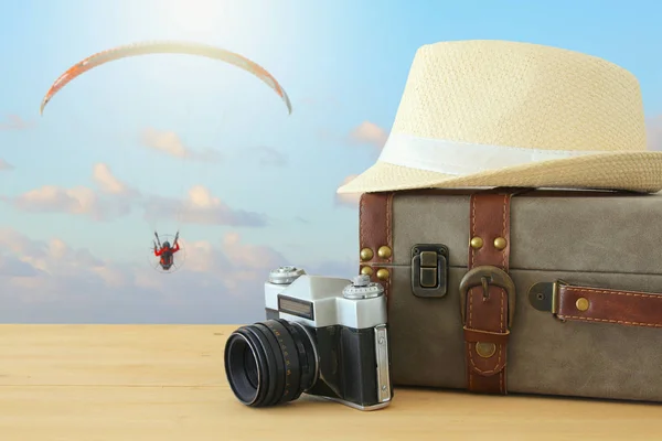 Equipaje vintage viajero, cámara y sombrero fedora sobre mesa de madera frente al cielo azul y paracaídas deporte. vacaciones y concepto de vacaciones . — Foto de Stock