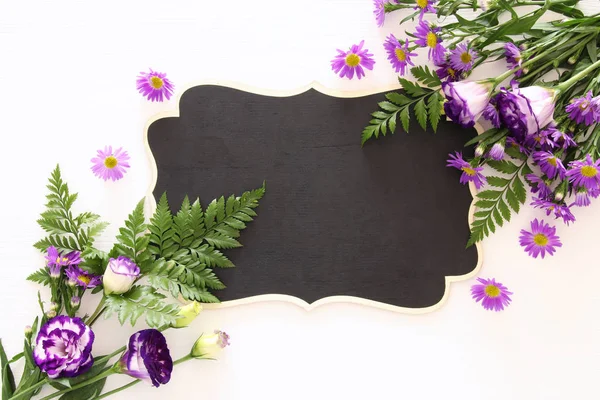 Ovanifrån av lila vackra blommor arrangemang och Tom blackboard över vit trä bakgrund. Kopiera utrymme. — Stockfoto