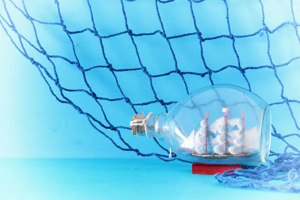 Nautische concept afbeelding met zeilboot in de fles en fishnet over blauwe houten tafel en achtergrond. Selectieve aandacht. — Stockfoto