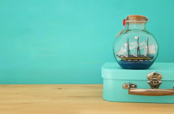 Nautical concept image with sail boat in the bottle and blue suitcase box over wooden table. Selective focus. — Stock Photo, Image