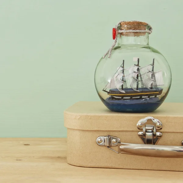 Nautical concept image with sail boat in the bottle and old suitcase box over wooden table. Selective focus. — Stock Photo, Image