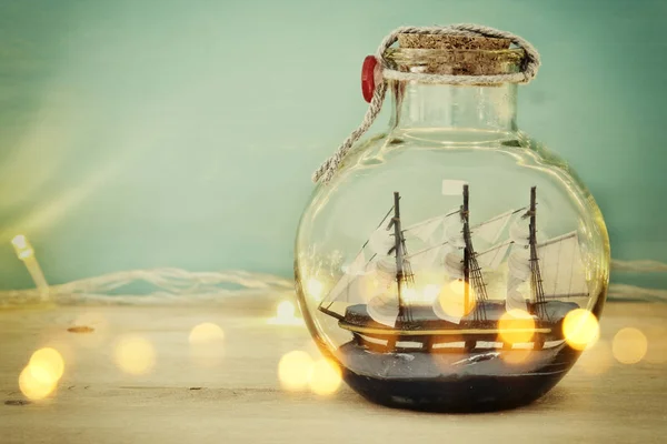 Concepto náutico imagen con barco de vela en la botella y luces de guirnalda de oro sobre mesa de madera. Enfoque selectivo . — Foto de Stock