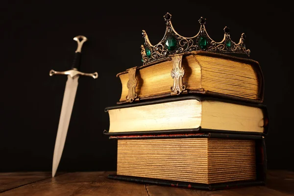 Imagen de llave baja de hermosa corona reina / rey sobre libro antiguo y espada. época medieval de fantasía. Enfoque selectivo —  Fotos de Stock