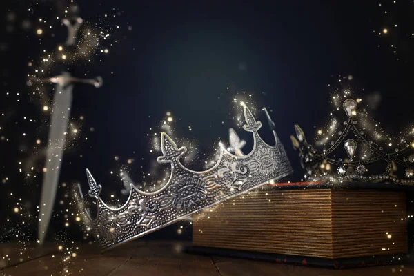 Laag sleutelbeeld van mooie koningin / koningskroon over antiek boek naast zwaard. fantasie middeleeuwse periode. Selectieve focus. Glitter fonkelende lichten — Stockfoto