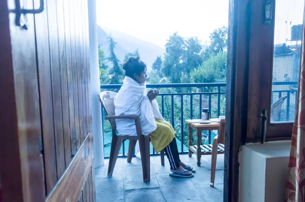 Eine junge frierende Touristin, die in Winterkleidung auf dem Balkon einer Ferienvilla sitzt und morgens eine heiße Tasse Tee in der Hand hält. Reisen Urlaub aktives Leben und gesunder Lebensstil Konzept. mou — Stockfoto