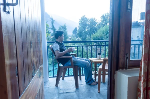 Reife erwachsene touristische Alleinreisende indischer Ethnie in Winterkleidung sitzen auf dem Balkon einer Ferienvilla und halten morgens eine heiße Tasse Tee vor dem Hintergrund der Bergstadt. Reisebüro — Stockfoto