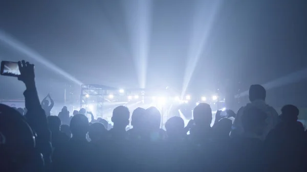 Silueta de gran audiencia multitud de personas felices de pie en el recinto del concierto aplaudiendo aplaudiendo el uso de teléfonos inteligentes levantando las manos en el aire frente a la banda de música disfrutando. Escenario reflejo de luz en frente . — Foto de Stock