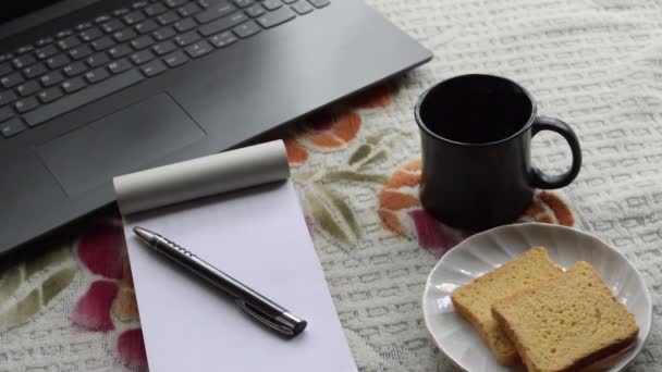 ノートパソコンの黒い色のペンと白の支配紙のノートブックで朝の日差しの中で朝食コーヒー トップオフィスの作業机の背景にセラミックカップソーサーとビスケット ライフスタイルイメージ — ストック動画