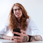 Lächelnd stylischer tätowierter Geschäftsmann mit lockigem Haar, Smartphone in der Hand