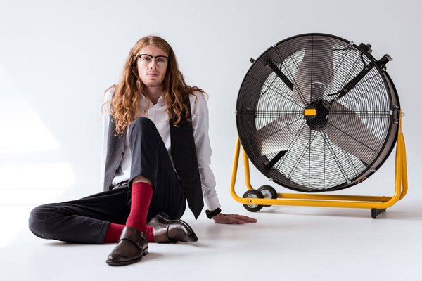 stylish businessman with curly hair sitting on floor near big fan