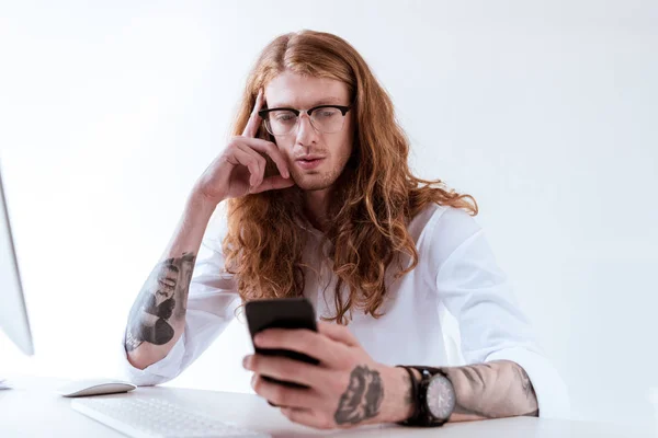 Stylish tattooed businessman with curly hair using smartphone at table in office — Stock Photo