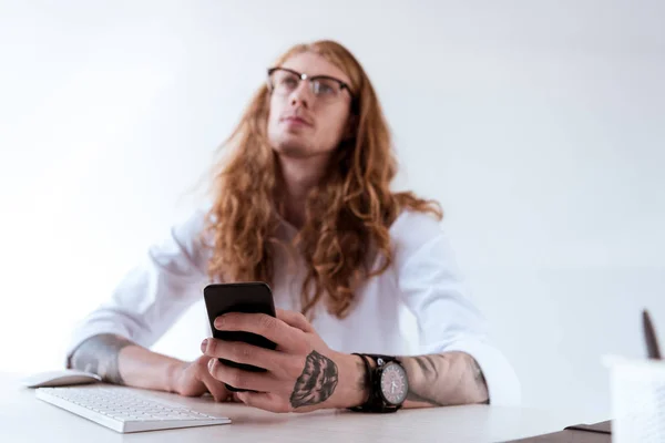 Pensive stylish tattooed businessman with curly hair holding smartphone and looking up — Stock Photo