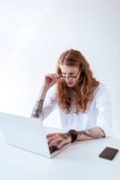 Homem de negócios tatuado elegante com cabelo encaracolado trabalhando com laptop no escritório e tocando óculos — Fotografia de Stock