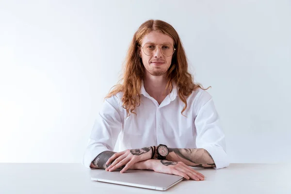 Стильный татуированный бизнесмен с вьющимися волосами сидит за столом с ноутбуком и смотрит на камеру, изолированную на белом — стоковое фото