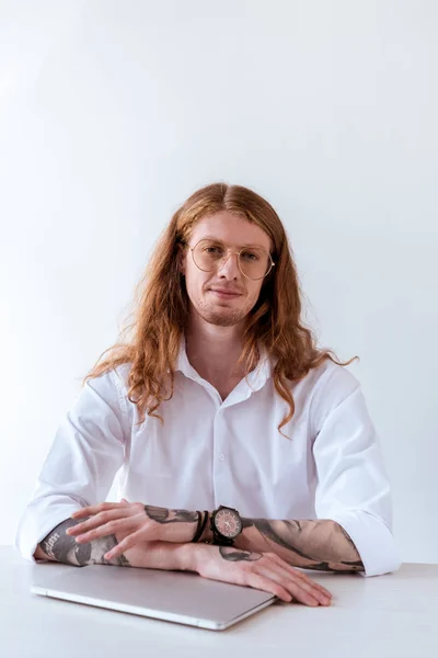 Homem de negócios tatuado elegante com cabelo encaracolado sentado à mesa com laptop e olhando para a câmera — Fotografia de Stock