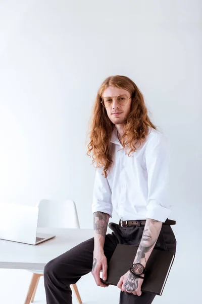Stylish tattooed businessman with curly hair sitting on table and looking at camera in office — Stock Photo