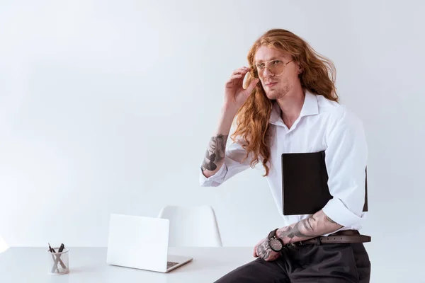 Stylischer tätowierter Geschäftsmann mit lockigem Haar, der auf dem Tisch sitzt und im Büro eine Brille anfasst — Stockfoto