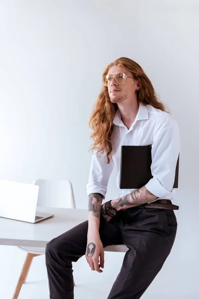 Elegante hombre de negocios tatuado con el pelo rizado sentado en la mesa y mirando hacia otro lado en la oficina - foto de stock