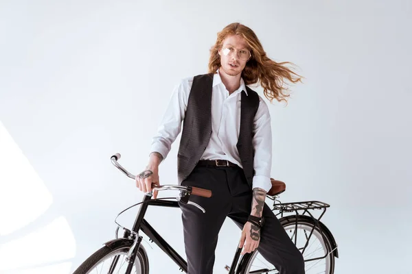 Stylish tattooed businessman with curly hair sitting on bicycle and looking at camera — Stock Photo