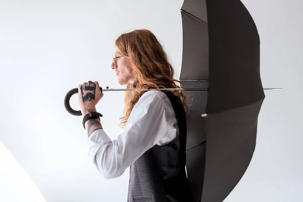 Vista lateral de homem de negócios tatuado elegante com cabelo encaracolado segurando guarda-chuva preto — Fotografia de Stock
