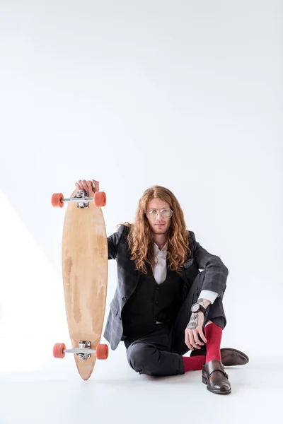 Stylish businessman with curly hair sitting on floor and holding skateboard — Stock Photo