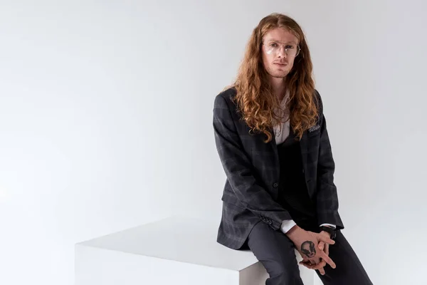 Stylish tattooed businessman with curly hair sitting on white cube isolated on white — Stock Photo