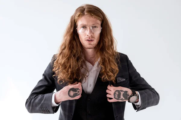 Elegante hombre de negocios tatuado con pelo rizado tocando la chaqueta y mirando a la cámara aislada en blanco - foto de stock