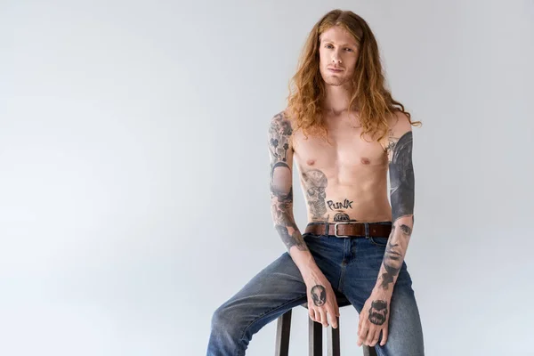 Bonito sem camisa tatuado homem com cabelos cacheados sentado na cadeira e olhando para a câmera isolada no branco — Fotografia de Stock