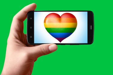 Telefon ekranında kalp şeklinde bir Lgbt bayrağı var. Eldeki akıllı telefon arka plandaki hromakey bayrağının kalbini gösteriyor. 