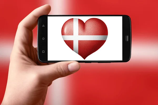 Danmark flagga i form av ett hjärta på telefonskärmen. Smartphone i handen visar ett hjärta flagga. — Stockfoto